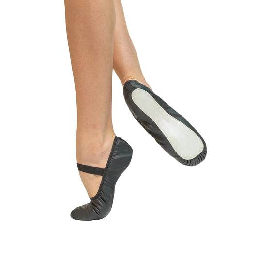 Black ballet slippers worn on a dancer's feet, shown against a plain light gray background. One slipper is on the ground and the other is lifted to show the sole.